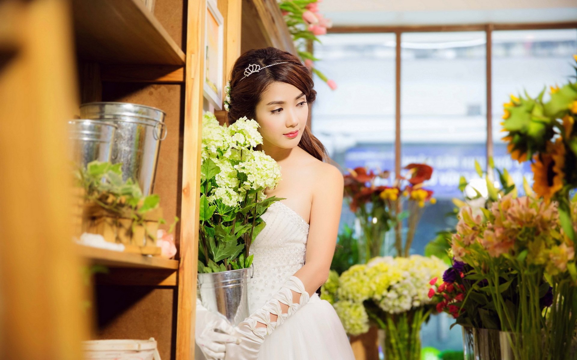 A woman named Linh Napie stands gracefully in a flower shop, wearing a white dress and holding a bouquet, surrounded by vibrant blooms. This HD wallpaper captures her elegance and beauty.