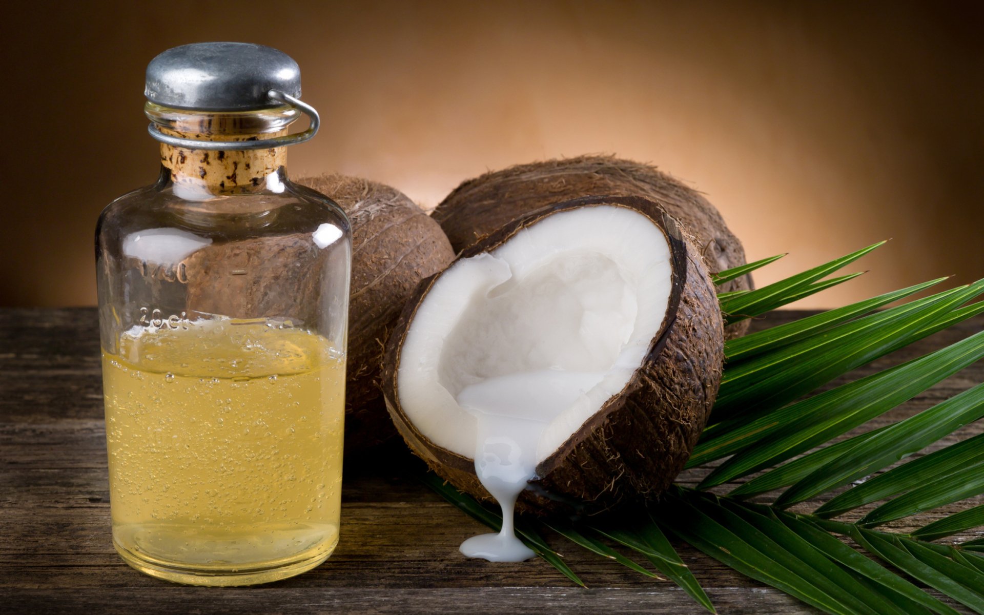 HD desktop wallpaper showing a man-made spa scene with a glass bottle of oil, a cracked coconut oozing milk, and a green palm leaf on a wooden surface.