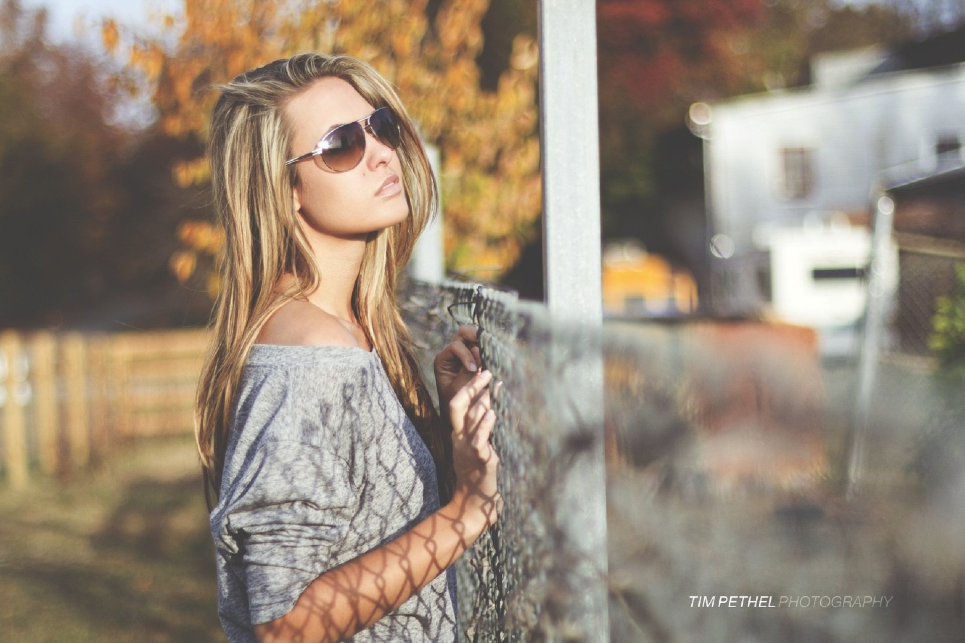 A woman wearing sunglasses leans on a fence with a thoughtful expression in a sunlit, autumn setting. HD desktop wallpaper and background.