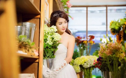 A woman named Linh Napie stands gracefully in a flower shop, wearing a white dress and holding a bouquet, surrounded by vibrant blooms. This HD wallpaper captures her elegance and beauty.