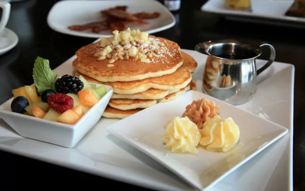 A stack of fluffy pancakes topped with nuts, accompanied by butter, fruit medley, and a small pot of syrup, captured in a vibrant 4K Ultra HD background.