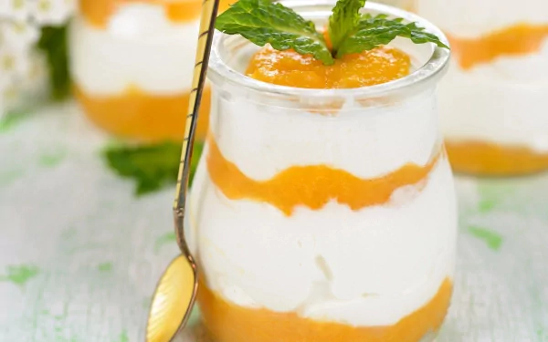A close-up 4K Ultra HD dessert wallpaper showing a glass filled with layered creamy white yogurt and bright orange fruit puree, garnished with fresh mint leaves and a spoon.