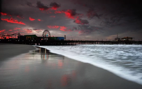 A stunning sunset over the ocean at Santa Monica Pier, featuring a Ferris wheel illuminated against a dramatic sky, with gentle waves lapping at the sandy beach, captured in HD.