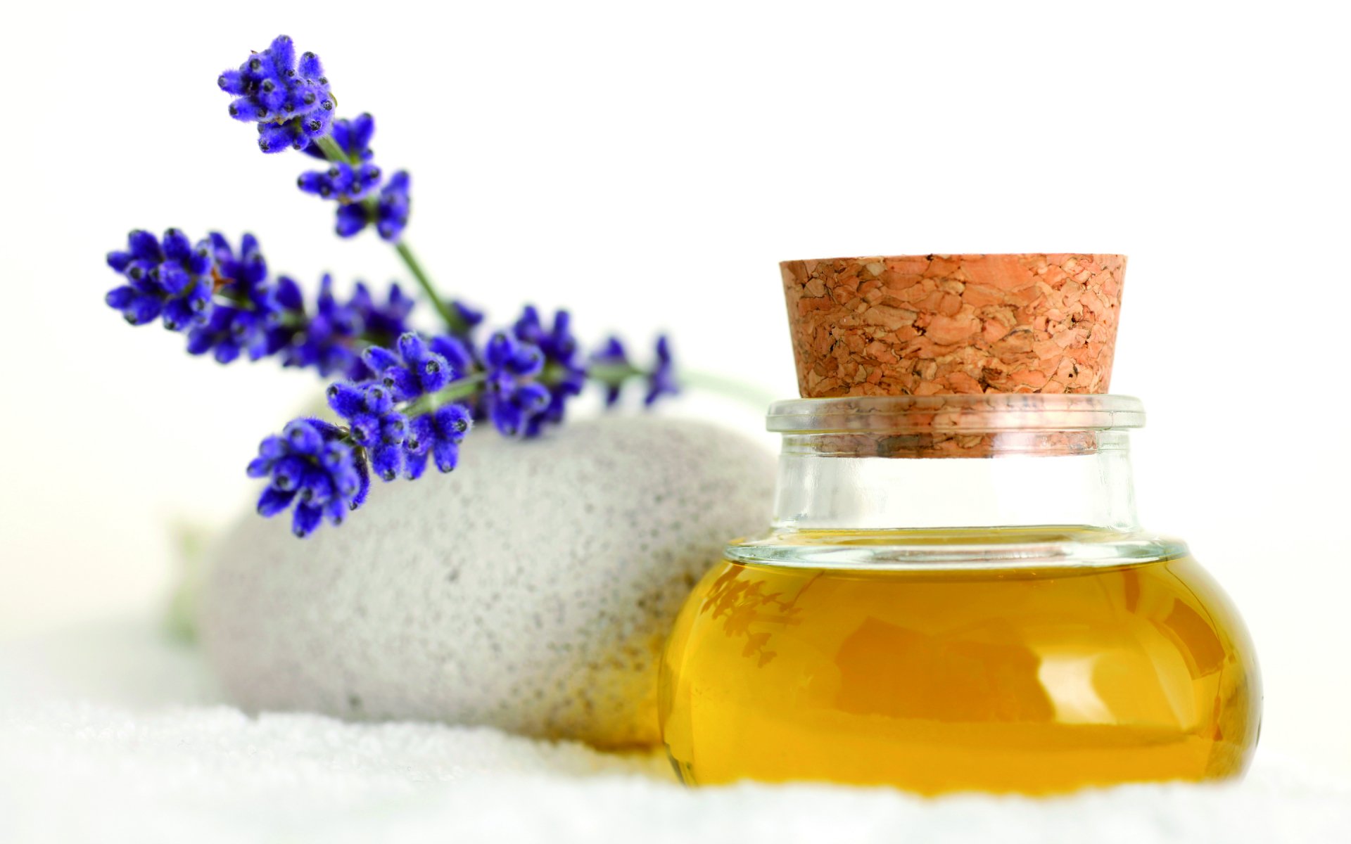 HD desktop wallpaper featuring a man-made spa scene with a small bottle of oil, lavender sprigs, and a smooth white stone on a soft white surface.