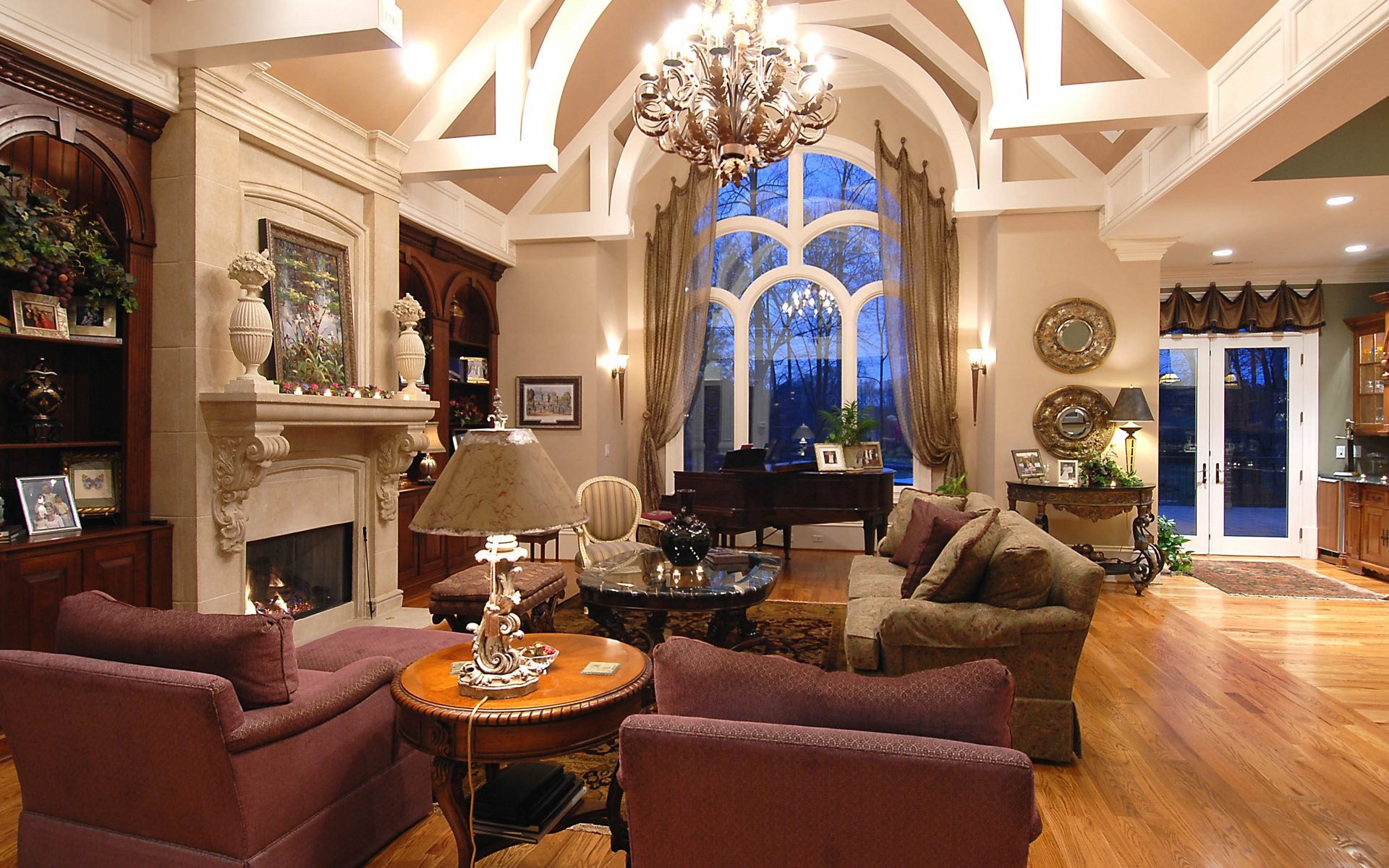 HD desktop wallpaper of an elegant, man-made living room featuring a grand chandelier, large arched window, cozy seating, and warm wooden flooring.