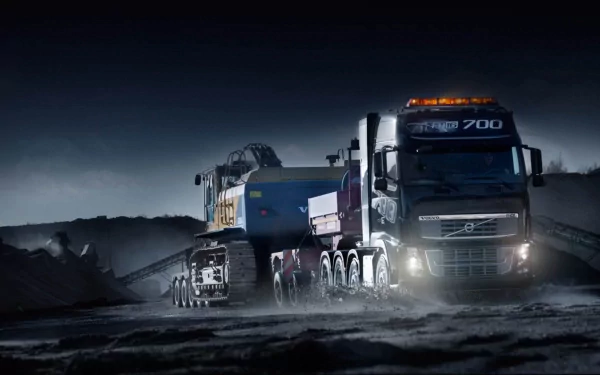 HD desktop wallpaper featuring a Volvo 700 truck hauling heavy machinery through a rugged, dark landscape at night.