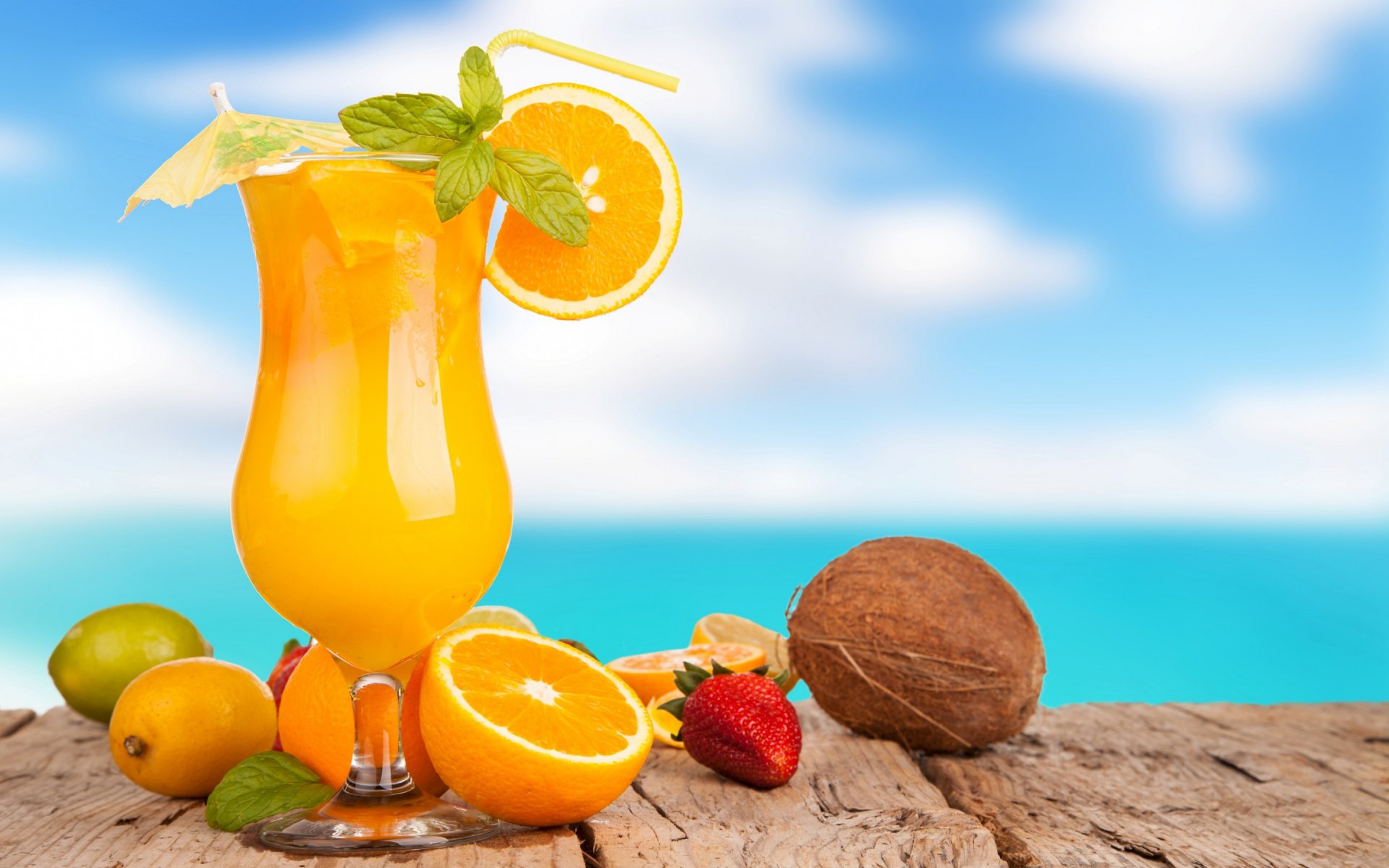 HD PC desktop wallpaper featuring a vibrant orange cocktail garnished with mint and citrus, set on a wooden surface with tropical fruits and a blurred ocean background.