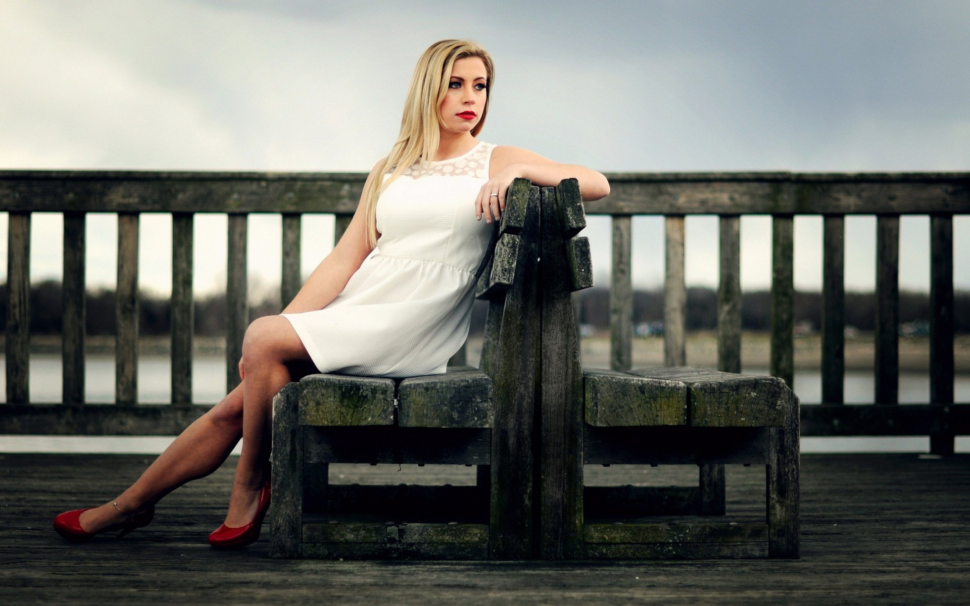 HD desktop wallpaper featuring a woman in a white dress and red shoes sitting on a wooden bench, exuding a contemplative mood against a moody sky.