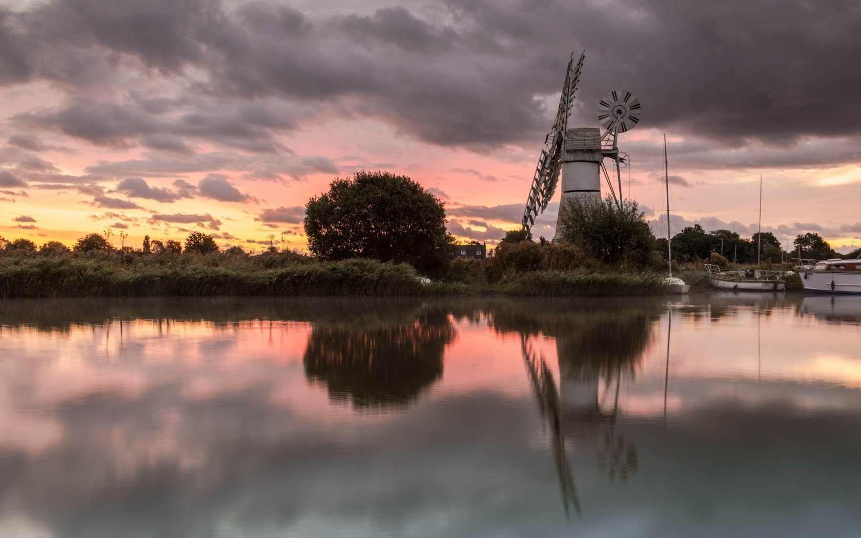 HD PC desktop wallpaper/background: man-made windmill by a calm reflective river at sunset, boats and trees silhouetted beneath dramatic, colorful clouds.