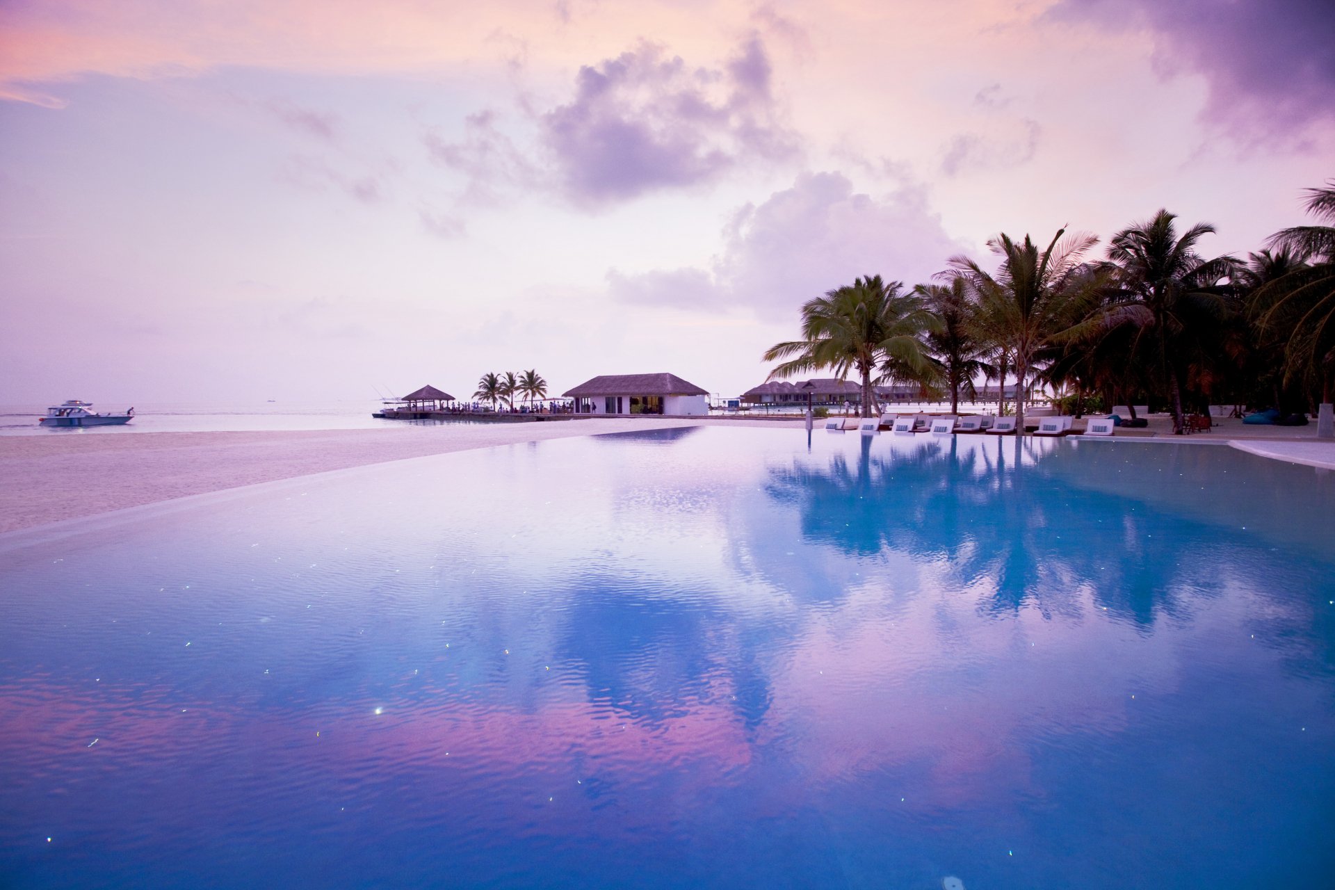 4K Ultra HD desktop wallpaper featuring a serene man-made pool with palm trees and a distant hut under a pastel purple sky by the water.