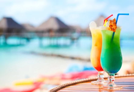HD PC desktop wallpaper featuring vibrant tropical cocktails on a table with a blurred beach and overwater bungalows in the background.