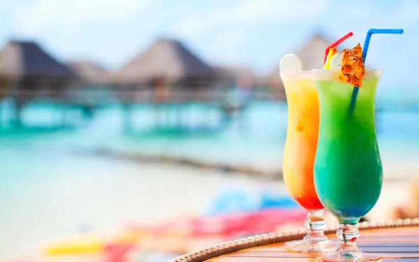 HD PC desktop wallpaper featuring vibrant tropical cocktails on a table with a blurred beach and overwater bungalows in the background.