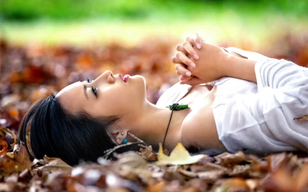 Asian woman with black hair lying down on a bed of leaves, wearing a necklace and earrings. The HD desktop wallpaper captures a peaceful moment in nature.