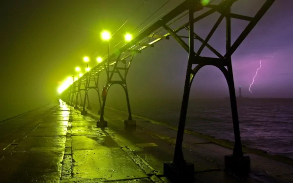 A dramatic HD wallpaper showcasing a lit bridge over water, illuminated by yellow lights amidst a stormy night, with lightning illuminating the sky in the background.