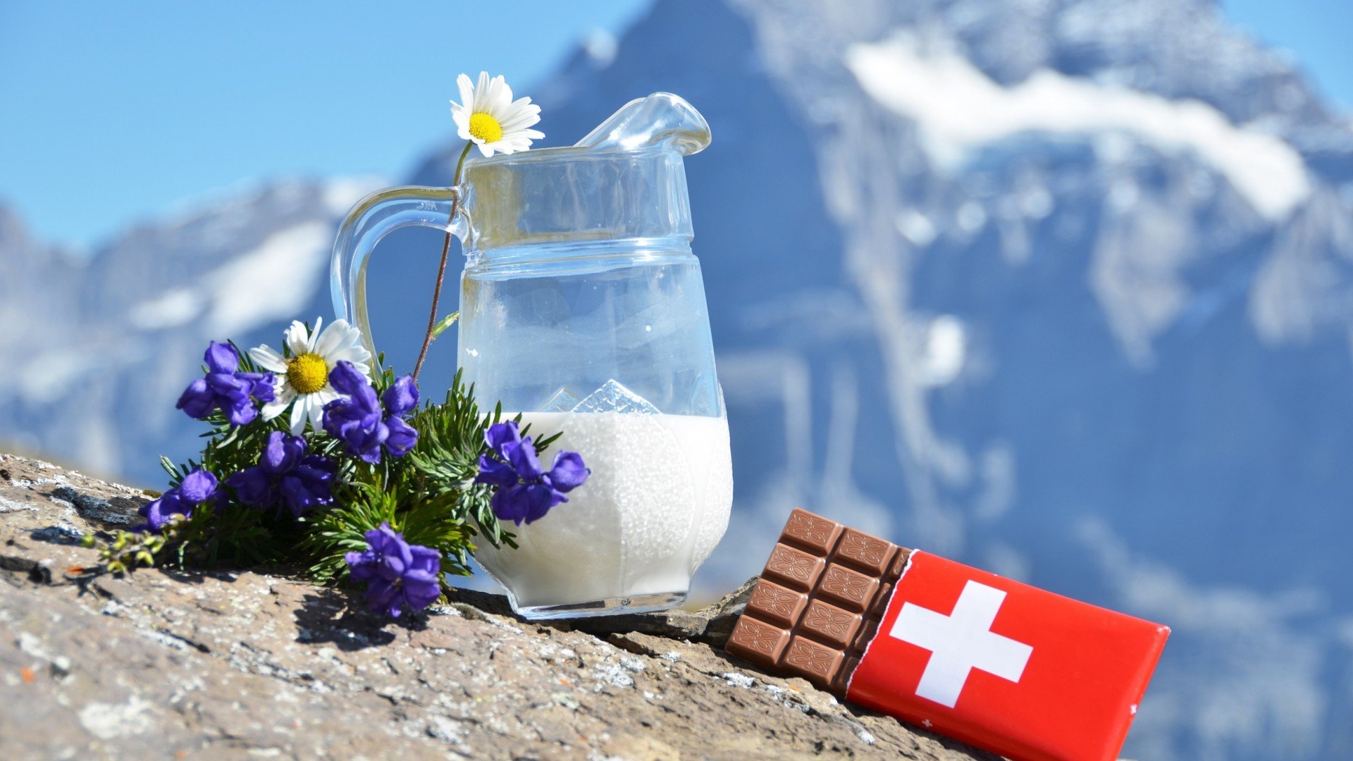 A picturesque scene featuring a jug of milk and Swiss chocolate against a backdrop of mountains, complemented by vibrant flowers, creating a delightful food-themed HD desktop wallpaper.