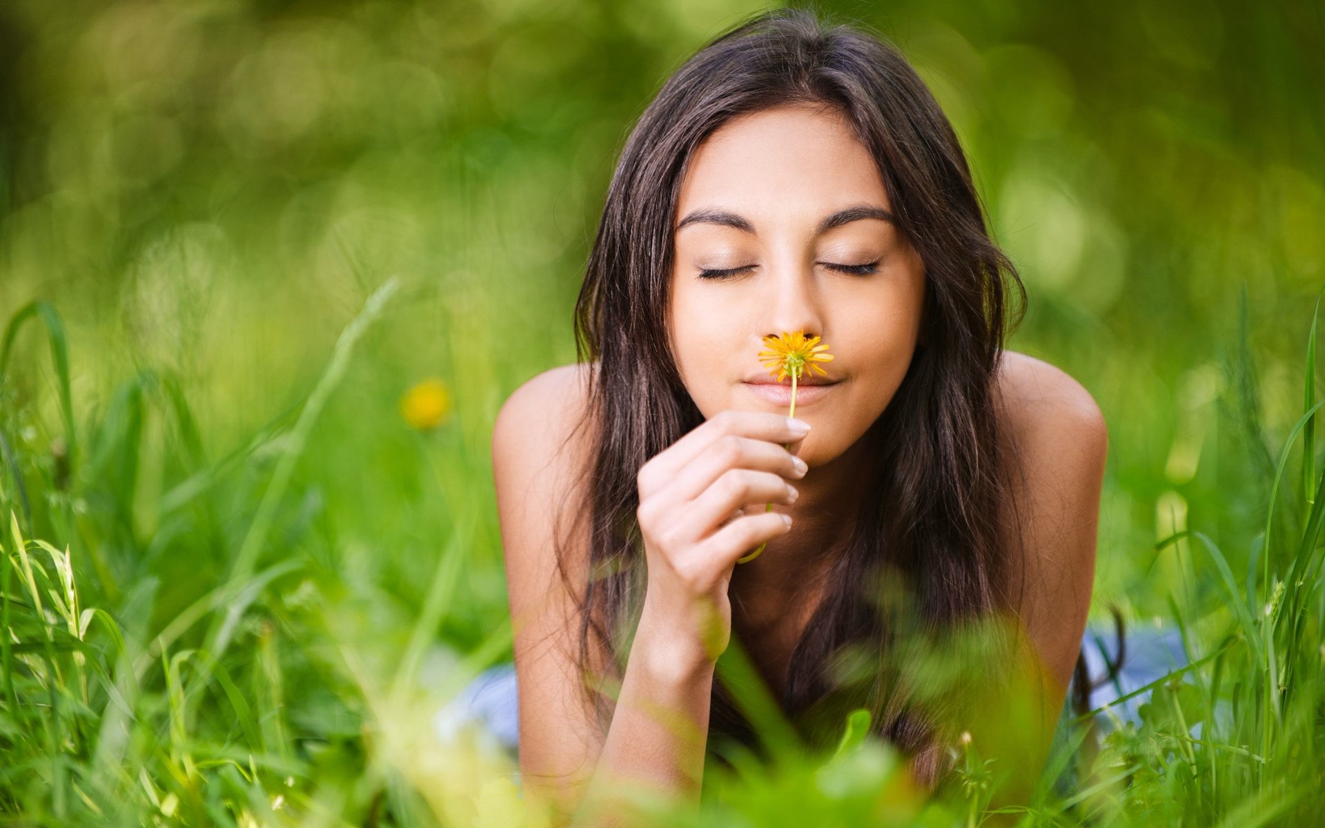 HD PC desktop wallpaper of a woman lying in green grass, eyes closed, gently holding and smelling a yellow flower, evoking a calm and peaceful mood.