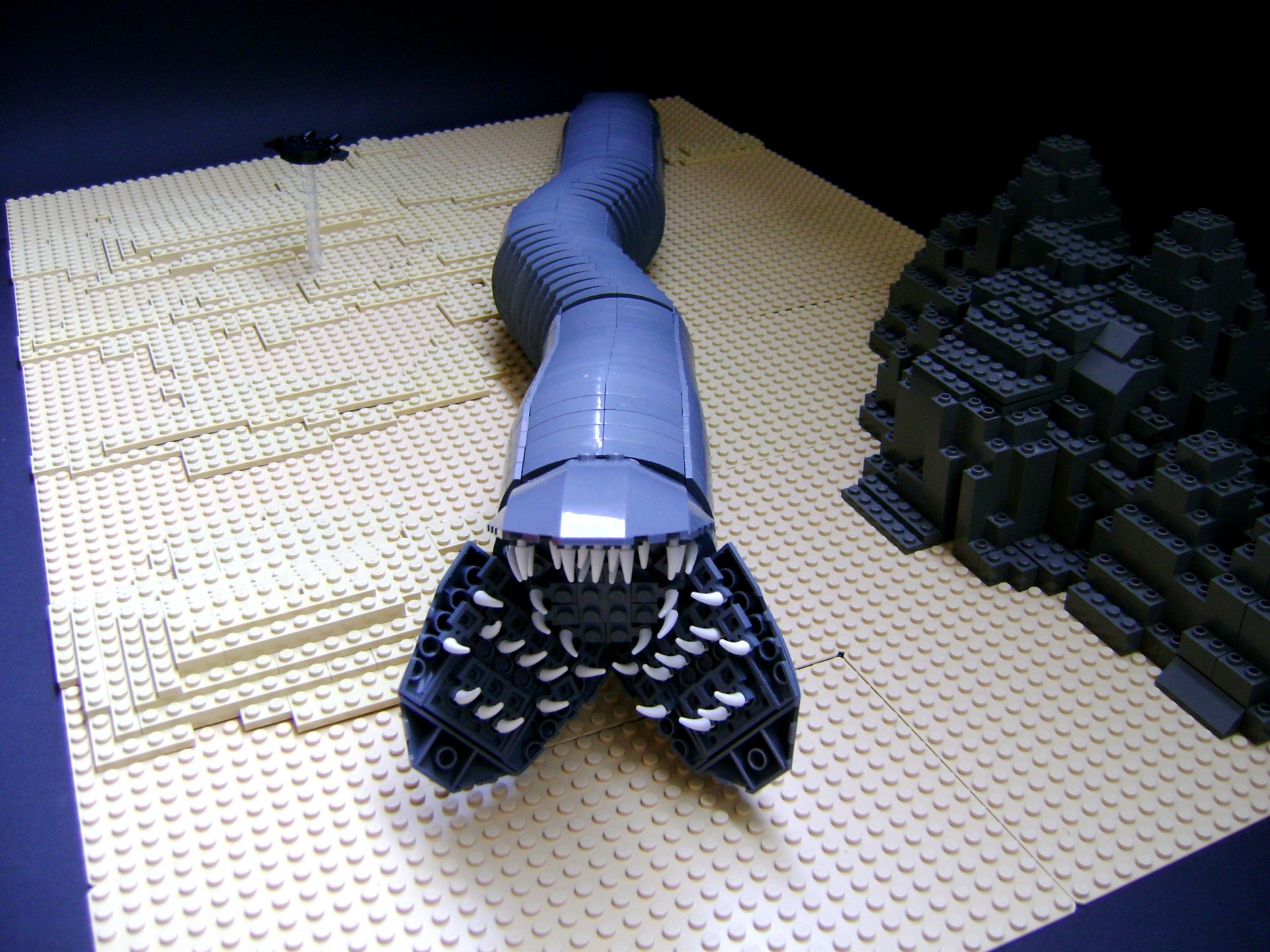 Sci-Fi Dune scene: LEGO sandworm emerging from tan desert beside a dark rock formation — 2K Quad HD PC desktop wallpaper and background.