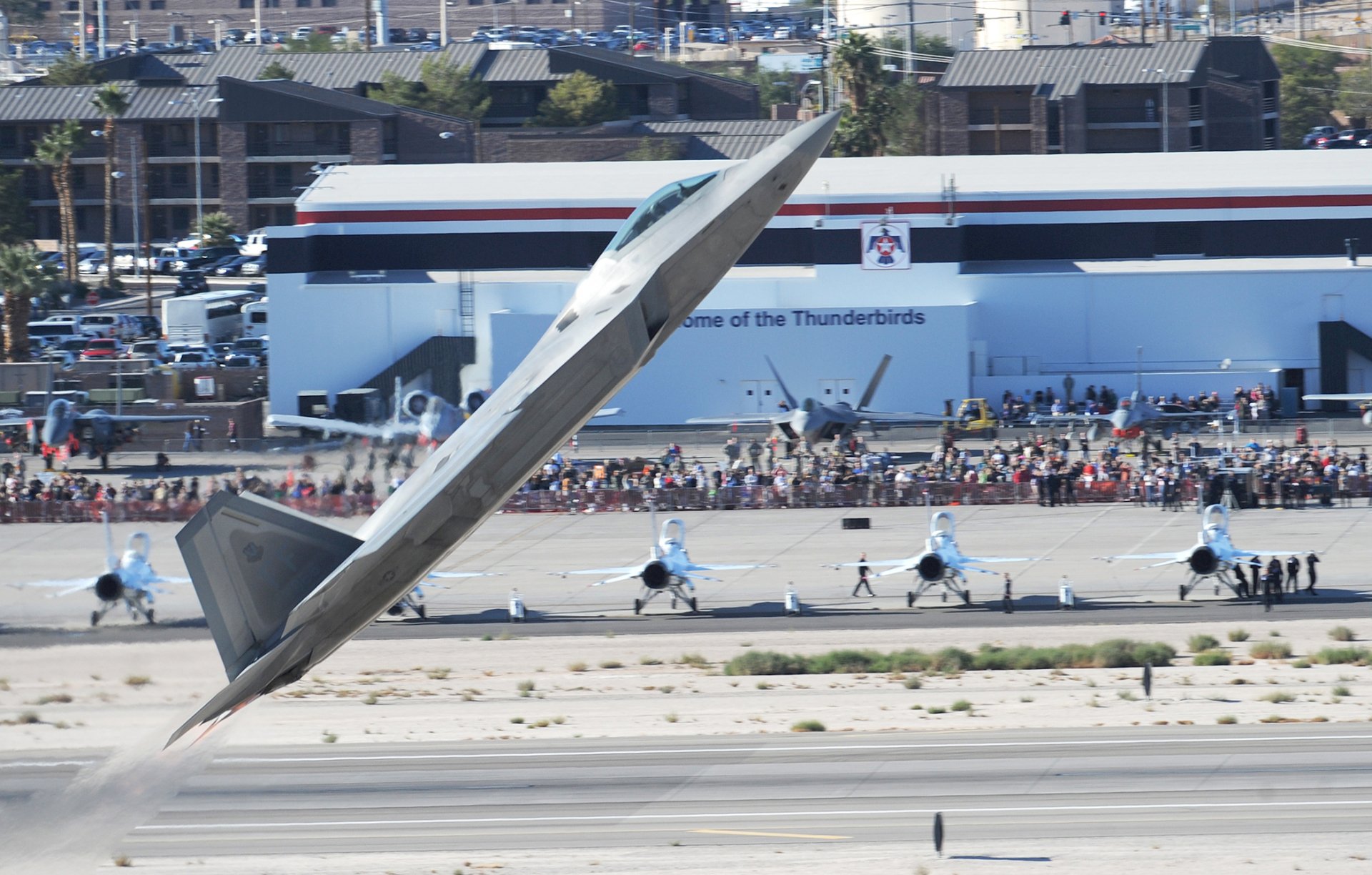 HD desktop wallpaper showcasing a Lockheed Martin F-22 Raptor military jet performing a sharp climb at an airshow with spectators and Thunderbirds jets in the background.