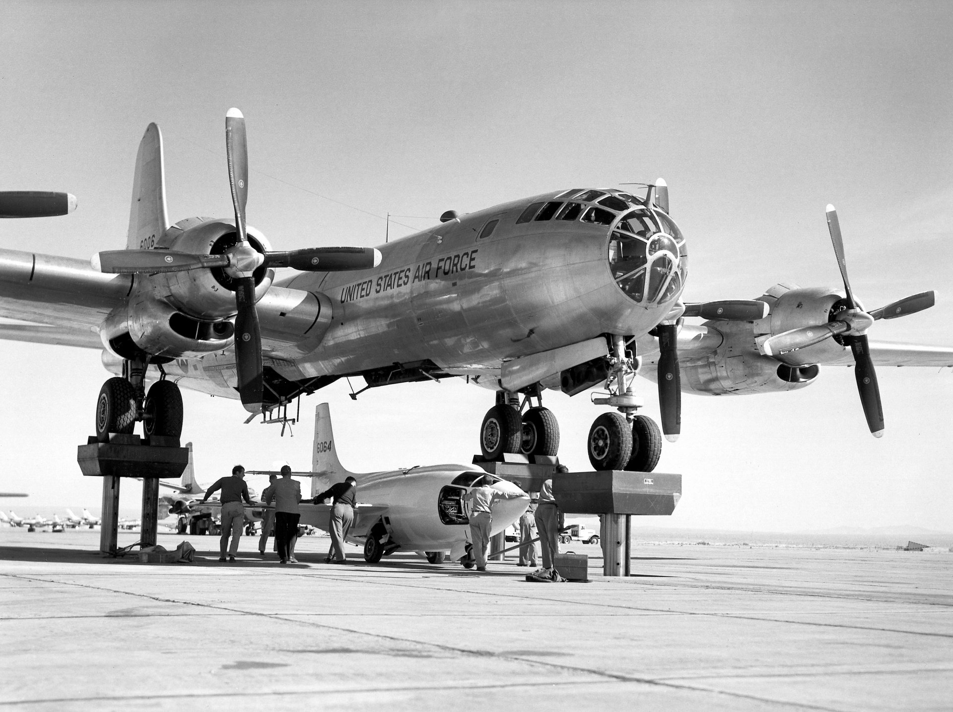 Black-and-white 2K Quad HD PC desktop wallpaper: Bell X-1 rocket plane tucked beneath a USAF bomber on the tarmac as crew work around the aircraft.