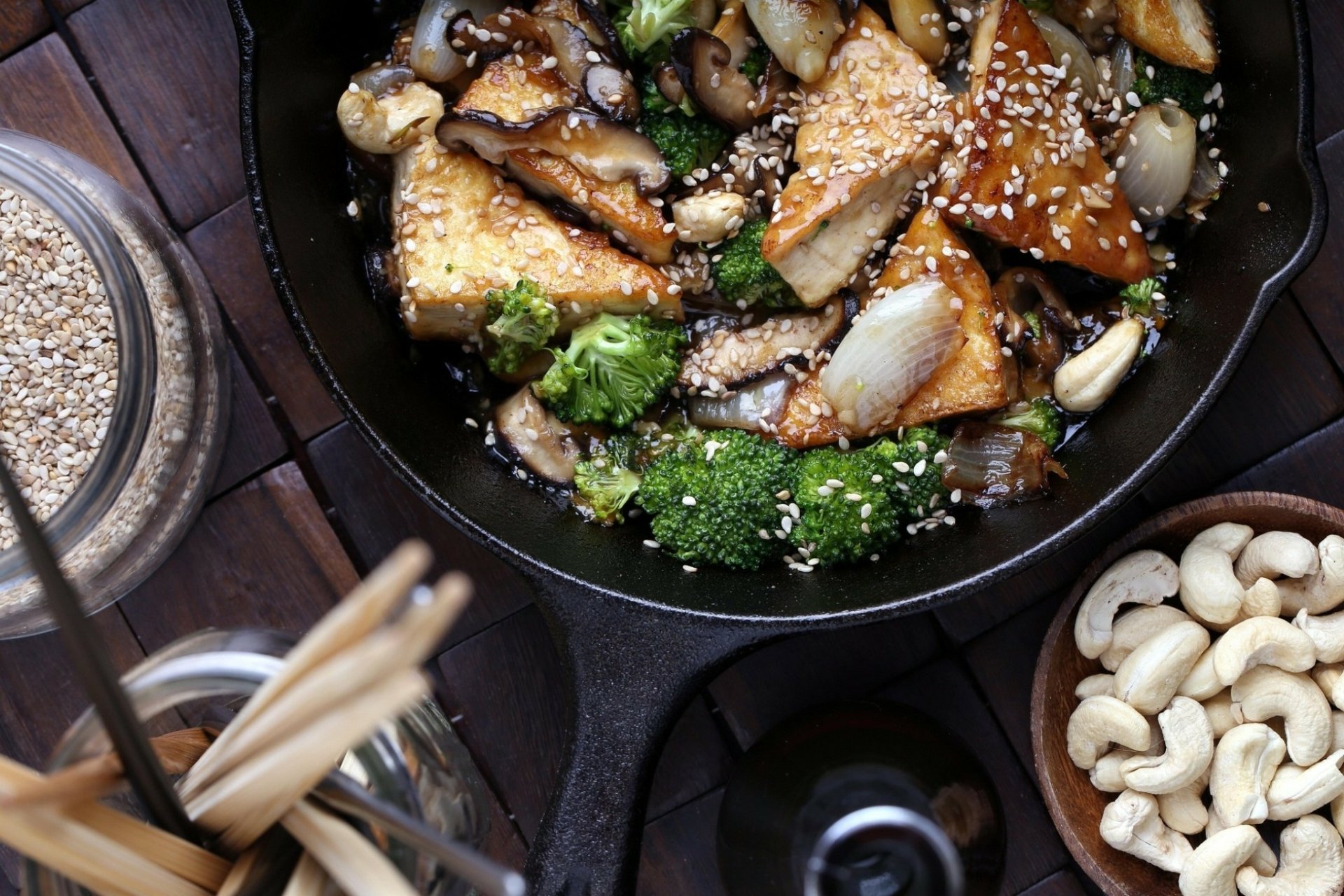 A vibrant skillet filled with seasoned tofu, broccoli, and mushrooms, garnished with sesame seeds, surrounded by cashews and chopsticks, making a striking food-themed HD desktop wallpaper.