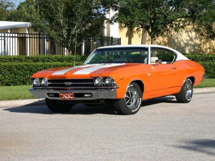 An orange Chevrolet Chevelle SS parked on a street, showcasing its classic muscle car design, vibrant color, and distinctive racing stripes against a backdrop of greenery.