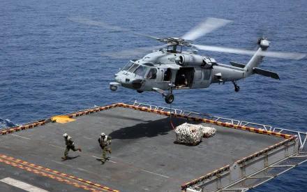 4K Ultra HD image of a military Sikorsky SH-60 Seahawk helicopter hovering above a ship deck over the ocean, with personnel managing cargo below.