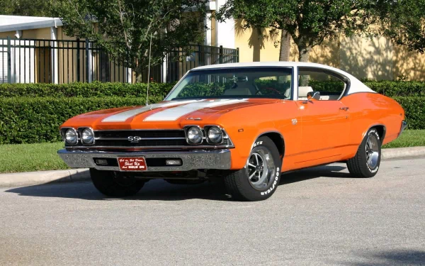 An orange Chevrolet Chevelle SS parked on a street, showcasing its classic muscle car design, vibrant color, and distinctive racing stripes against a backdrop of greenery.