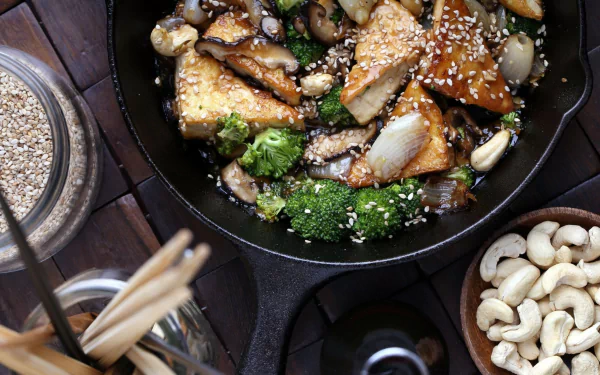 A vibrant skillet filled with seasoned tofu, broccoli, and mushrooms, garnished with sesame seeds, surrounded by cashews and chopsticks, making a striking food-themed HD desktop wallpaper.