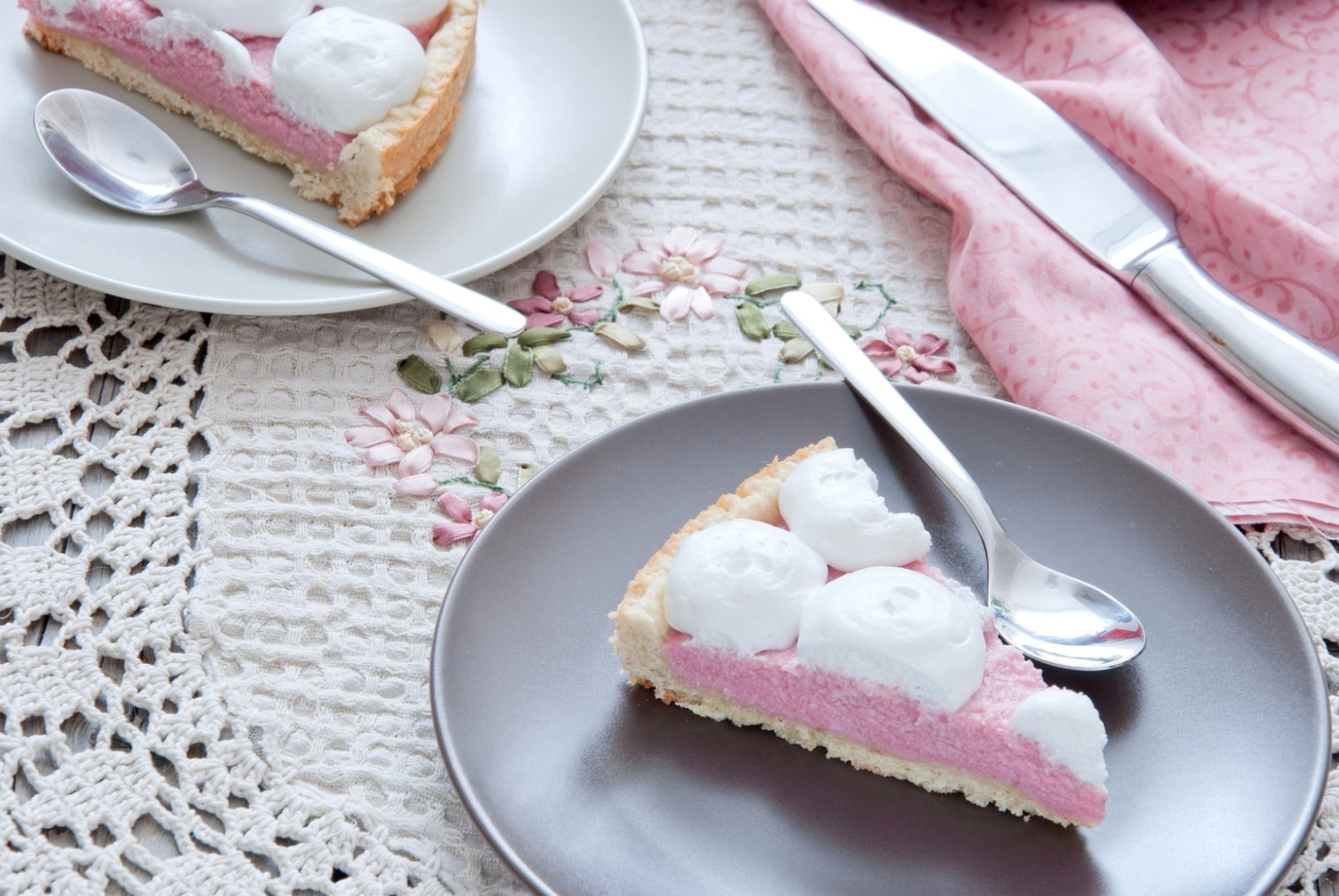Two slices of pink cake topped with whipped cream on elegant plates, set on a lace tablecloth with a pink cloth napkin and utensils, creating a charming food-themed desktop wallpaper.