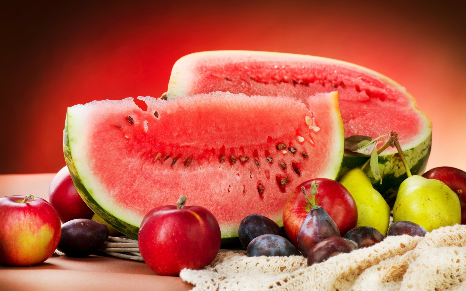 A vibrant HD desktop wallpaper featuring fresh fruit, including a sliced watermelon, red apples, and various plums, set against a warm, colorful background.