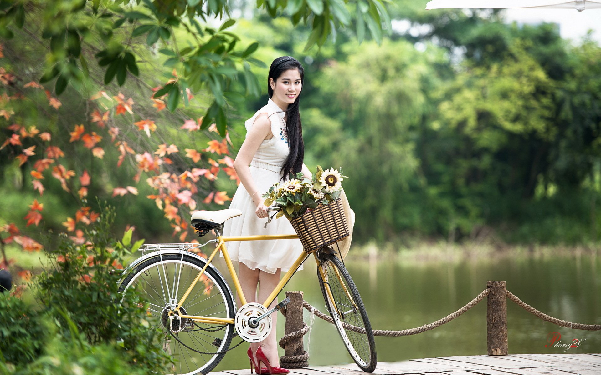 A woman stands beside a yellow bicycle adorned with flowers, set against a serene lakeside backdrop and lush greenery. This HD image makes a vibrant desktop wallpaper.