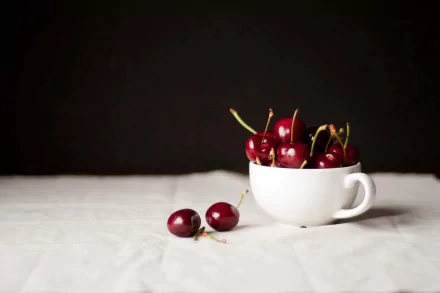 HD desktop wallpaper featuring fresh cherries in a white cup on a white cloth with a dark background, highlighting vibrant red fruit and simple elegance.