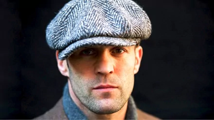HD desktop wallpaper featuring celebrity Jason Statham wearing a herringbone flat cap, with a stern expression against a dark background.