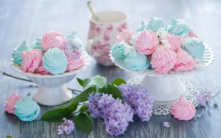 A vibrant HD desktop wallpaper featuring pastel pink and blue meringues arranged on elegant stands, accompanied by lilac flowers and a decorative mug, creating a delightful foodie scene.