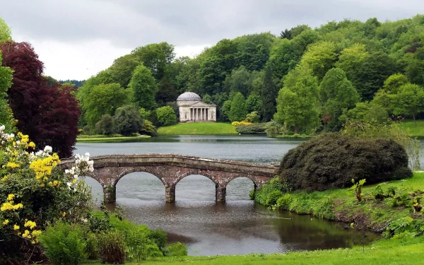 A serene landscape featuring a stone bridge over a lake, surrounded by dense forest, flowering shrubs, a green field, and a classical monument near the water's edge.