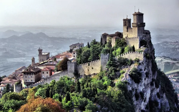 HD desktop wallpaper featuring the historic man-made fortress and towers of San Marino perched atop a rocky hill, surrounded by lush greenery and distant mountains.