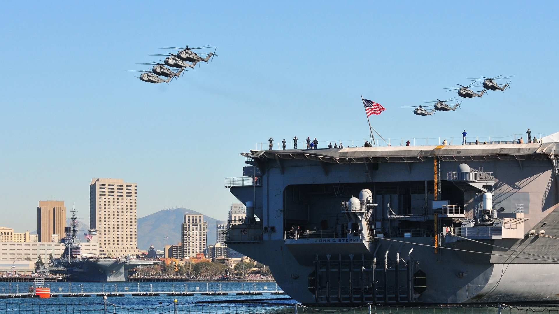 Military Sikorsky CH-53E Super Stallion helicopters fly in formation above a naval ship with an American flag, set against a city skyline in this HD desktop wallpaper.