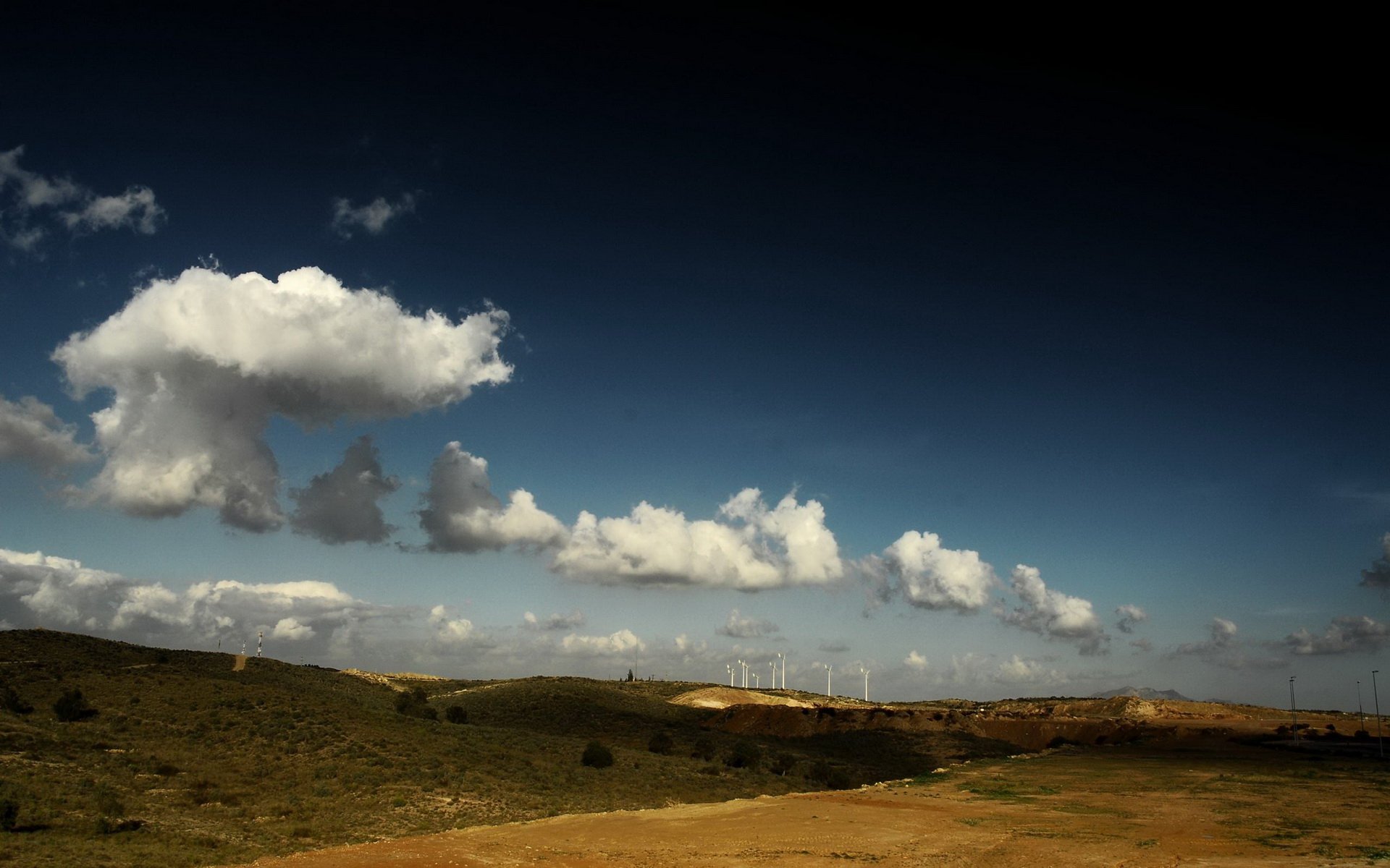 HD PC desktop wallpaper and background showing a nature landscape: rolling grass and dirt path leading to distant wind turbines beneath a deep blue sky dotted with white cumulus clouds.