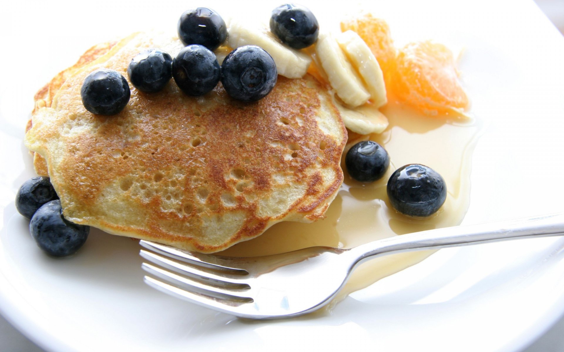 HD PC desktop wallpaper featuring a close-up of a golden pancake topped with blueberries, banana slices, and syrup on a white plate with a fork.