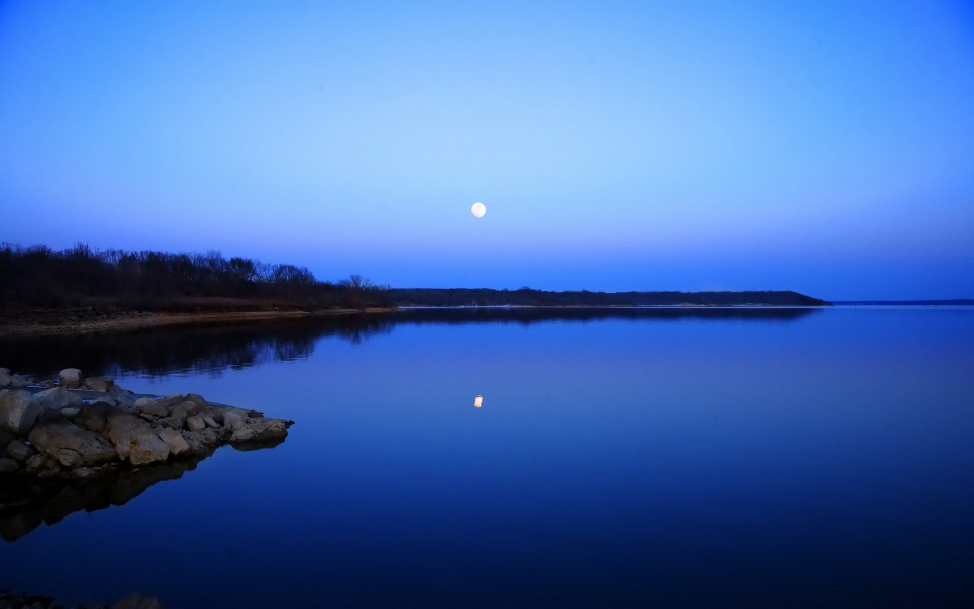 HD PC desktop wallpaper — nature scene with a full moon over a calm blue lake at twilight, moonlight reflecting on the water, rocky foreground and distant shoreline