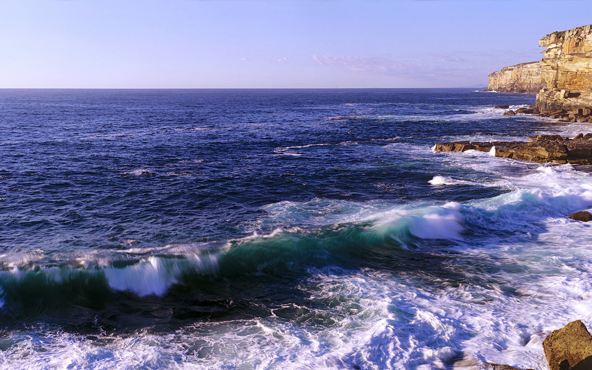 HD PC desktop wallpaper showing a vibrant coastline with waves crashing against rocky cliffs under a clear blue sky.