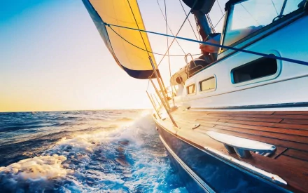A sleek sailboat cuts through vibrant blue waters, illuminated by the warm glow of the sunset, creating a stunning HD desktop wallpaper and background.