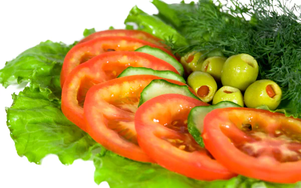 A vibrant salad featuring sliced tomatoes, cucumbers, green olives, and fresh greens, set against a clean white background. This HD desktop wallpaper celebrates fresh, healthy food.