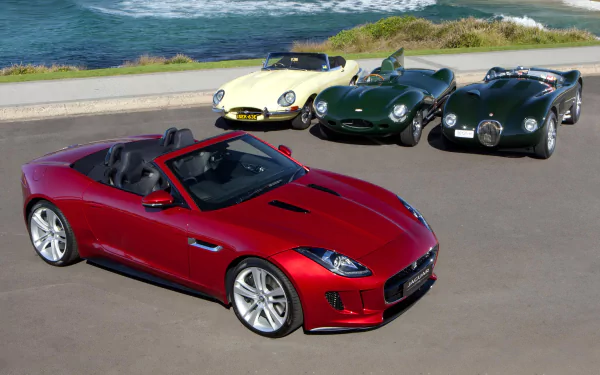 A HD desktop wallpaper featuring four Jaguar cars, including a red modern convertible and three classic Jaguar E-Type vehicles, parked near a coastal road with ocean views.