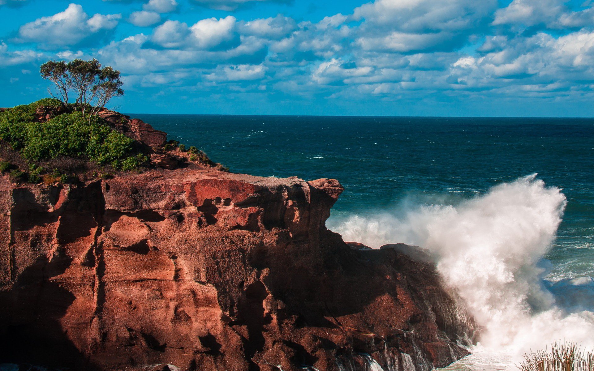 HD PC desktop wallpaper showcasing a rugged cliff with greenery under a blue sky, waves crashing against the rocks in a vibrant nature scene.