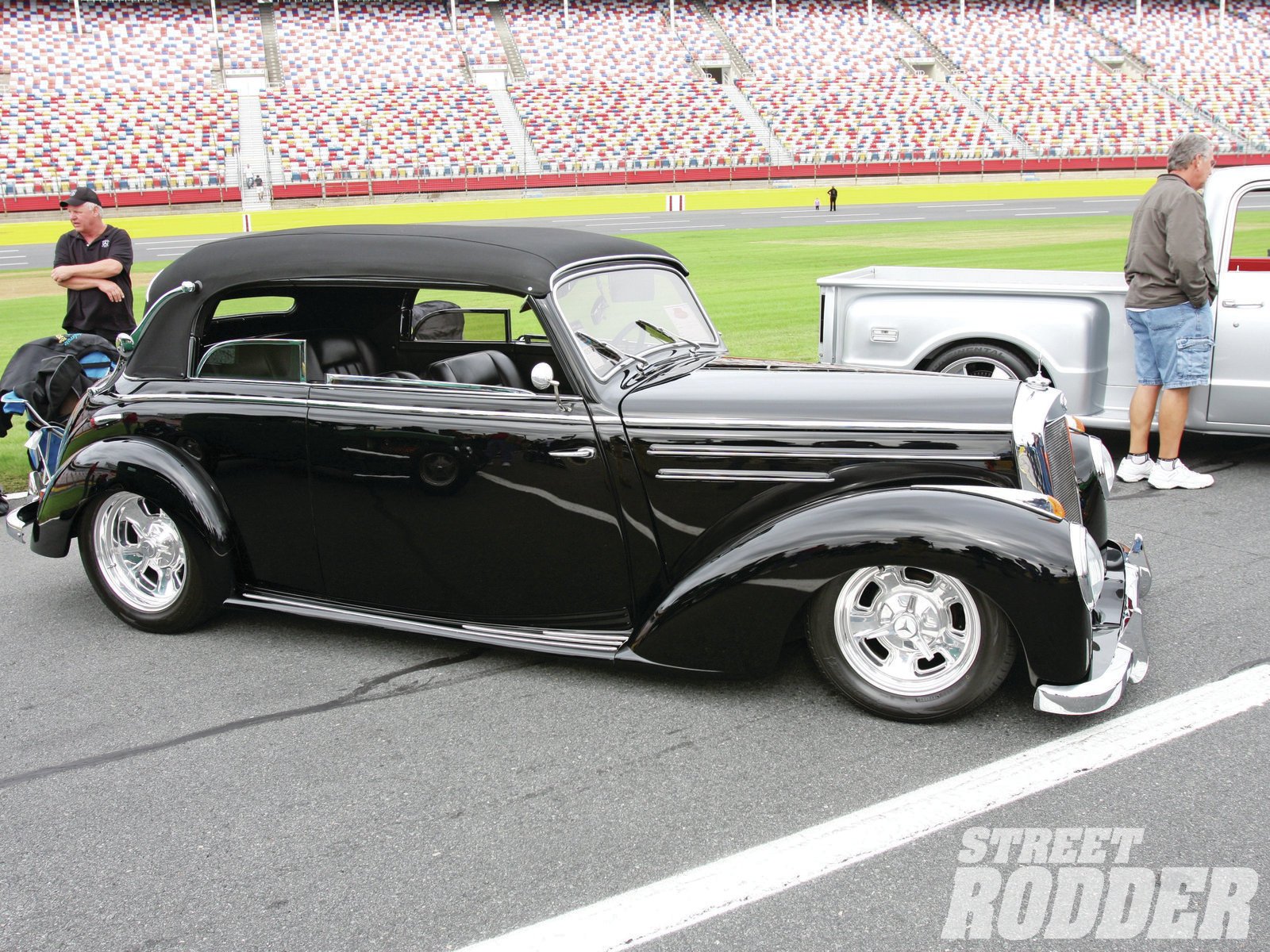 HD PC desktop wallpaper: glossy black vintage Mercedes‑Benz vehicle convertible with chrome wheels parked on a racetrack, spectators and another classic car behind.