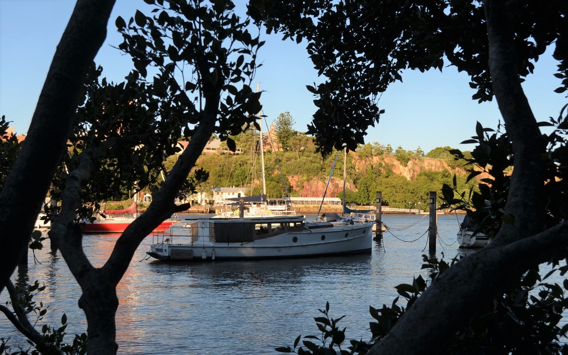 HD desktop wallpaper: boat vehicle moored on the Brisbane River, Australia, framed by silhouetted trees, with other vessels and riverbank homes in the background.