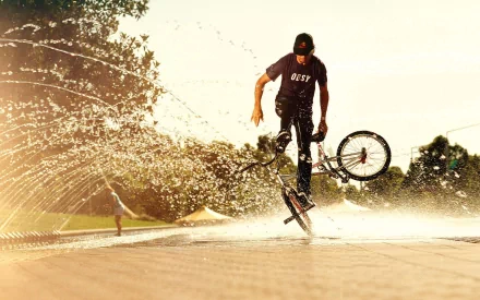 A man performs a bicycle trick through a water fountain in a park. The image is a high-definition desktop wallpaper and background, capturing a dynamic and energetic mood.