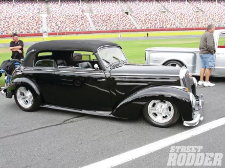 HD PC desktop wallpaper: glossy black vintage Mercedes‑Benz vehicle convertible with chrome wheels parked on a racetrack, spectators and another classic car behind.