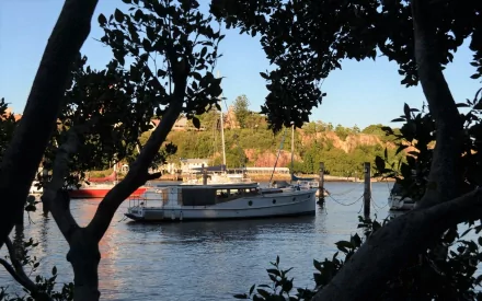HD desktop wallpaper: boat vehicle moored on the Brisbane River, Australia, framed by silhouetted trees, with other vessels and riverbank homes in the background.